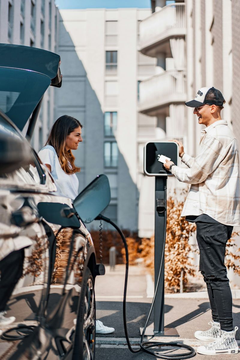 A man and a woman standing beside their black electric car while it charges at a kerbside post