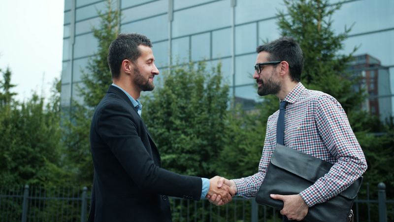 Two men shaking hands outdoors, one holding a document folder