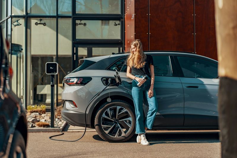 A woman leaning against her grey electric car with a coffee while it charges